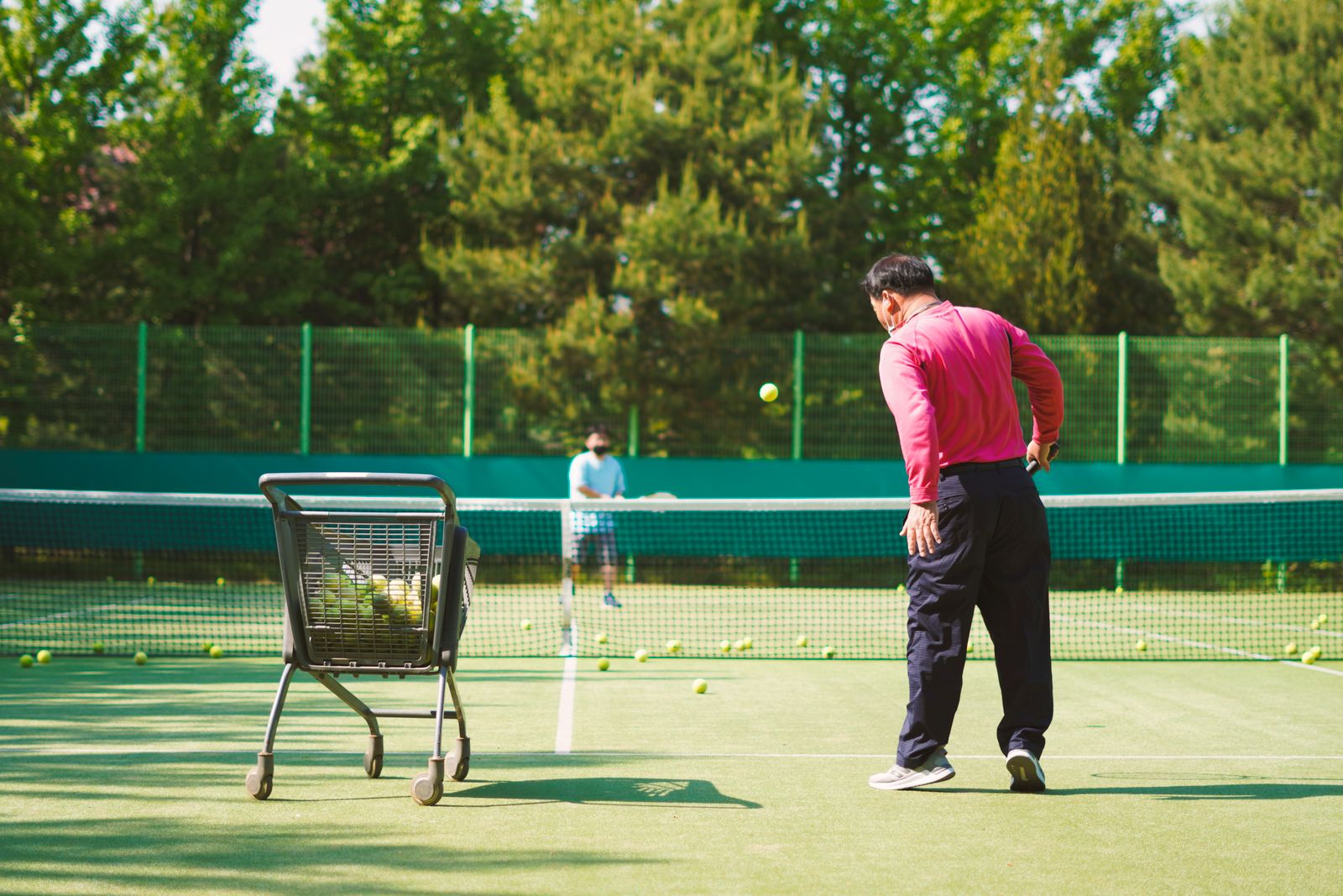 Two adults practicing tennis beside a ball cart on a court.